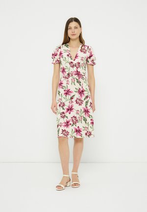 Kobieta stoi, ubrana w białą sukienkę do kolan z różowo-zielonym kwiatowym wzorem, bufkami, dekoltem w serek i białe sandały na obcasie.