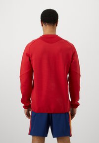 Κόκκινο μακρυμάνικο φούτερ με στρογγυλή λαιμόκοψη και ριχτές λεπτομέρειες, συνδυασμένο με μπλε σορτσάκι που έχει κόκκινες και κίτρινες λωρίδες στο πλάι.