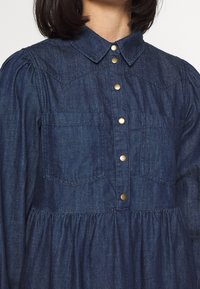 Tummansininen farkkimekko, jossa on napitettu etuosa, kaulus ja kaksi rintataskua. Pöhnätyt hihat ja kerätty yksityiskohta vyötäröllä.