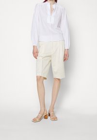 Valkoinen, pitkähihainen paita mandariinikauluksella, yhdistettynä vaaleanbeigeihin pliseerattuihin shortseihin ja kermaisiin sandaaleihin, joissa on paksu korko.