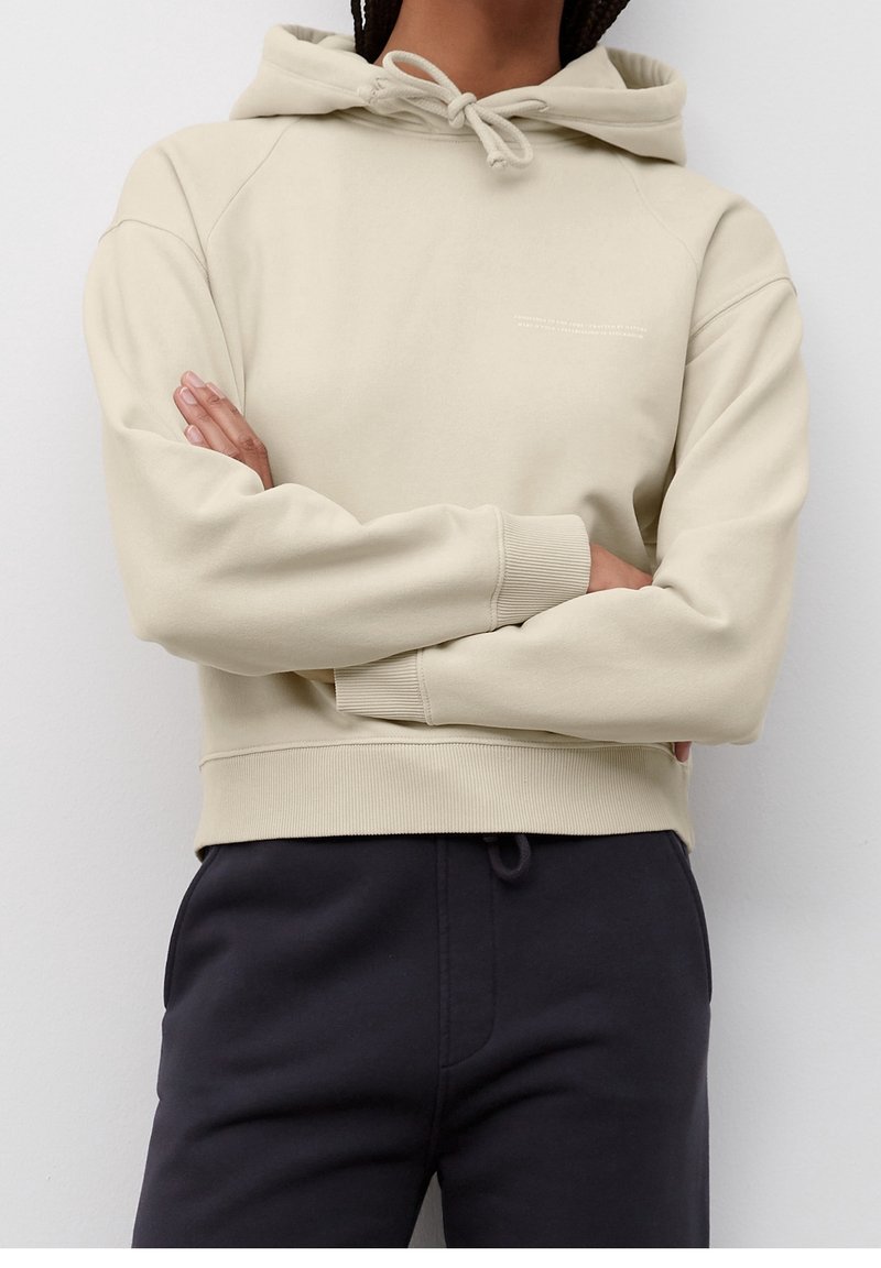 Personne debout, les bras croisés, portant un sweat à capuche beige clair avec la capuche relevée et des cordons, et un pantalon foncé, sur un fond uni.