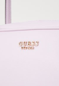 Bolso sintético lila con el logo dorado en relieve "GUESS" y detalle "EST 1981". Textura suave y elementos de diseño simples y modernos.