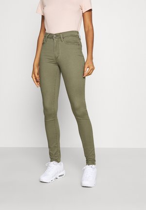 Mujer con jeans ajustados de talle alto en verde oliva, camiseta de manga corta rosa pálido y zapatillas blancas, de pie contra un fondo claro y sencillo.