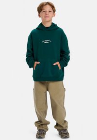 Πράσινο φούτερ με κουκούλα και μπροστινή τσέπη, με λογότυπο, συνδυασμένο με χακί φαρδιά παντελόνια και sneakers με σχέδια. Ομαλή υφή υφάσματος.