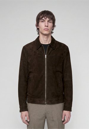 Young man wearing a dark brown suede zip-up jacket and checkered pants, standing against a plain light gray background.