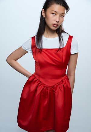 Mlajša ženska z dolgimi temnimi lasmi, oblečena v rdečo satensko obleko brez rokavov preko bele majice s kratkimi rokavi, stoji z roko na boku.