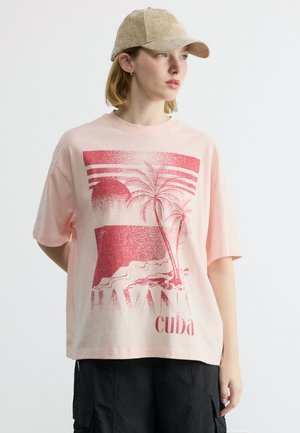 Νεαρό άτομο που φοράει μπεζ καπέλο και ροζ φαρδιά μπλούζα με κόκκινο γραφικό φοίνικα Havana Cuba και μαύρες βερμούδες, κοιτάζοντας δεξιά.