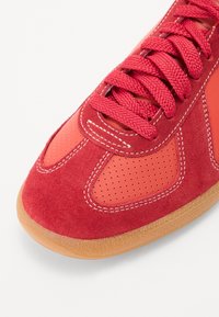 Chaussure de sport rouge en mélange de daim et de matériaux lisses, avec panneaux perforés, lacets assortis et semelle en caoutchouc gomme.