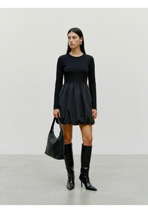 Woman wearing a black long-sleeve dress, black knee-high boots, and holding a black textured handbag against a plain light background.
