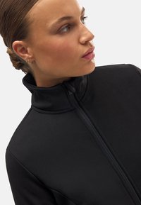 Chaqueta negra con cremallera y cuello alto, hecha de material suave. Presenta una vista lateral, mostrando un diseño elegante y un sutil detalle de textura.