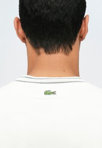 Vue arrière d'une personne avec des cheveux courts et noirs portant une chemise blanche avec un petit logo de crocodile vert près du col.