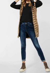 Vadderad guldväst med huva över en svart långärmad tröja, kombinerad med mörkblå åtsittande jeans och bruna högtoppsskor.