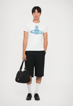 Witte T-shirt met een blauw grafisch ontwerp, gecombineerd met zwarte shorts, witte sokken en zwarte instapschoenen, terwijl er een zwarte tas wordt vastgehouden.