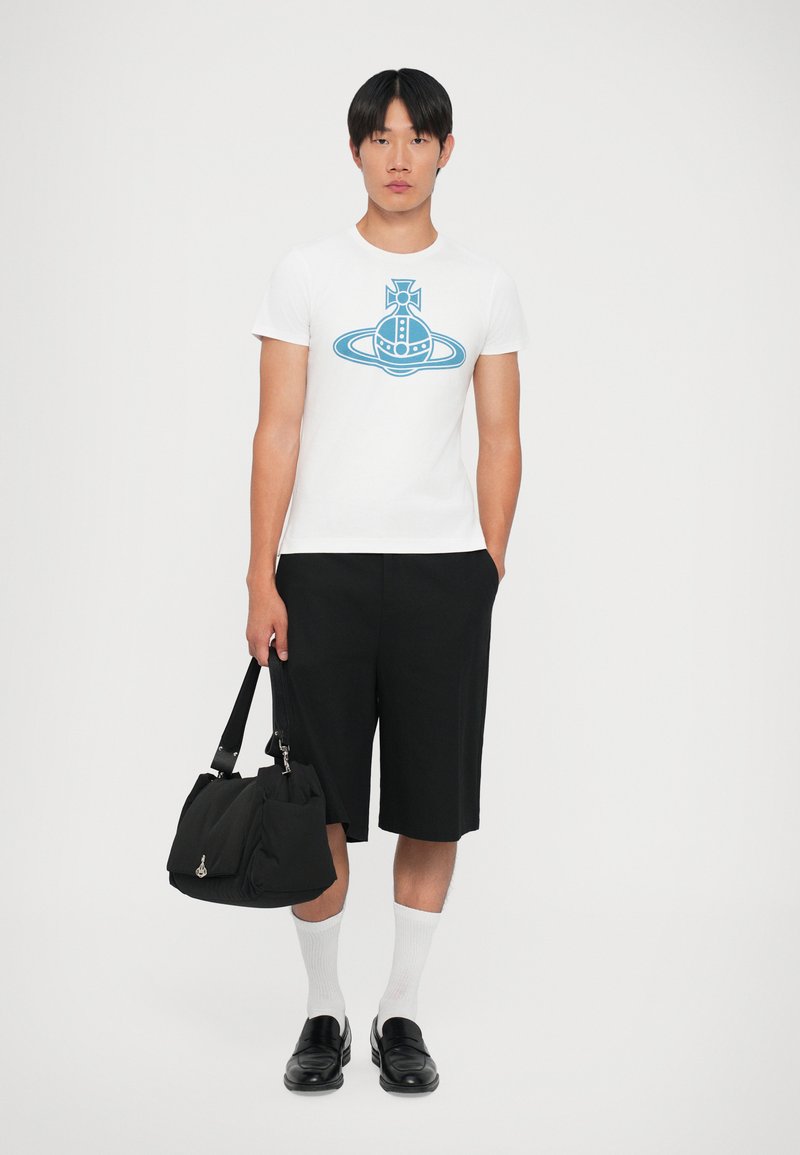 T-shirt blanche avec un design graphique bleu, associé à un short noir, des chaussettes blanches et des mocassins noirs, tout en tenant un sac noir.
