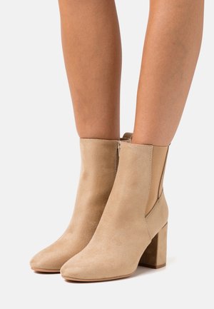 Beige suede ankle boots with a pointed toe, side elastic panels, and a chunky heel. Smooth texture, minimalistic design.