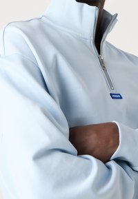 Sudadera de cremallera azul claro con un cuello alto, puños acanalados y una pequeña etiqueta de logo azul. Tela suave con una textura lisa.