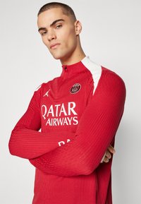 Piros sportos, hosszú ujjú felső, texturált anyagból, magas nyakkal és cipzárral. Fehér kiegészítőkkel és a "QATAR AIRWAYS" logóval az elején.