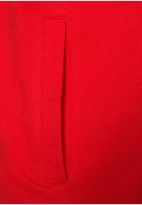 Roter Sweatshirtstoff mit glatter Textur, ausgestattet mit einer vertikalen Eingrifftasche auf der linken Seite, sorgfältig genähten Kanten und gleichmäßiger Farbe.