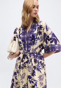 Γυναίκα φορώντας κρέμ φόρεμα με μπλε λουλουδάτα σχέδια, κρατώντας μια λευκή τσάντα, κοιτάζοντας στο πλάι μπροστά σε ένα απλό φόντο.