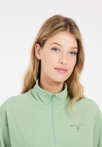 Světle zelený, žebrovaný top na půl zip s polo límečkem. Obsahuje malé vyšívané logo na hrudi. Měkká textura a ležérní design.
