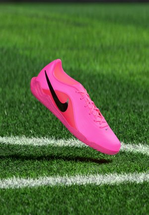 Pantof de fotbal Nike roz aprins cu swoosh negru plutind deasupra gazonului verde lângă liniile albe ale terenului.
