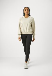 Beige fleece genser med ribbede mansjetter og nederkant, kombinert med svarte leggings og lyse sneakers, presentert på en modell mot en ensfarget bakgrunn.