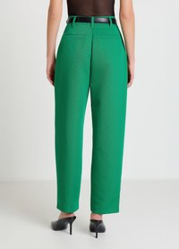 Pantalon vert taille haute avec deux poches arrière, texture lisse, et coupe jambes larges. Associé à une ceinture noire et des chaussures à talons.