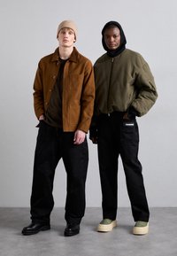 Dos individuos llevan prendas exteriores distintas: una chaqueta marrón con cuello, una bomber de color oliva oscuro y pantalones negros. El calzado incluye zapatos negros y zapatillas de plataforma verdes.