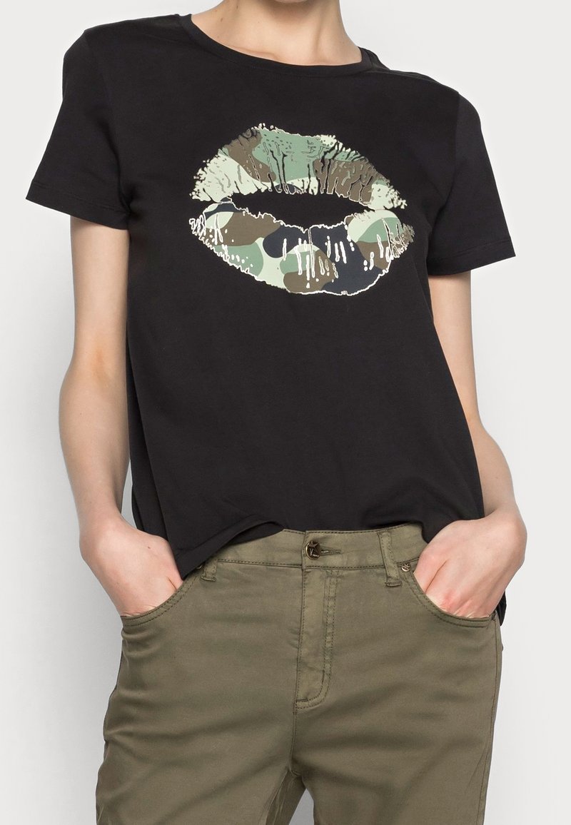 Svart bomulls t-shirt med en stor grafik av läppar i kamouflage-mönster. Kort ärm, rund halsringning, avslappnad passform. Kombinerad med olivgröna byxor.