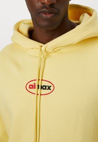 Yellow hoodie made of soft fabric, featuring a front zipper and a circular logo with "ail max" in black and red. Close-up view.