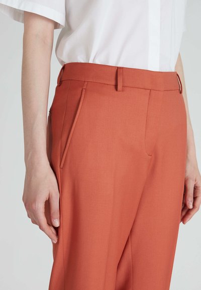 Pantaloni terracotta con una texture liscia, dotati di tasche laterali oblique e una vestibilità sartoriale. Abbinati a una camicia bianca a maniche corte.
