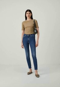 Kvinne som står iført en beige kortermet topp, blå smale jeans, brune leiligheter og en liten mørkegrønn skulderveske.