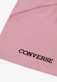 Šviesiai rožinė medvilninė marškinė su šonkaulinės tekstūros apdaila ir juodu "CONVERSE" logotipu, spausdintu šalia apatinio krašto. Paprastas, klasikinis dizainas.