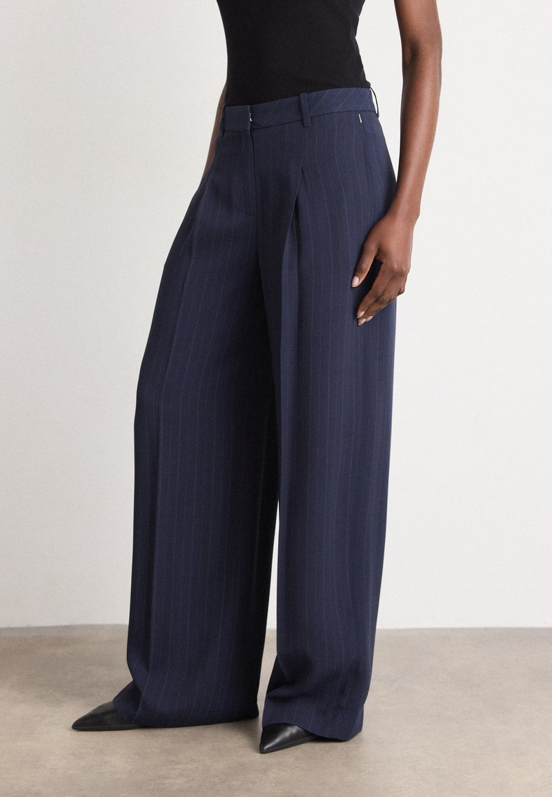 Femme portant un pantalon large à rayures bleu marine et des chaussures noires à bouts pointus, debout devant un fond clair uni.