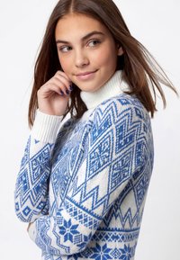 Mėlynas ir baltas megztinis su aukštu kaklu, geometriniais raštais ir ribotu rankogaliu. Minkšta tekstūra ir jauki išvaizda.