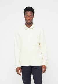 Ανοιχτόκίτρινο πουκάμισο με κουμπιά από υφασμάτινη υφή, με γιακά και μία τσέπη στο στήθος, συνδυασμένο με σκούρο παντελόνι.