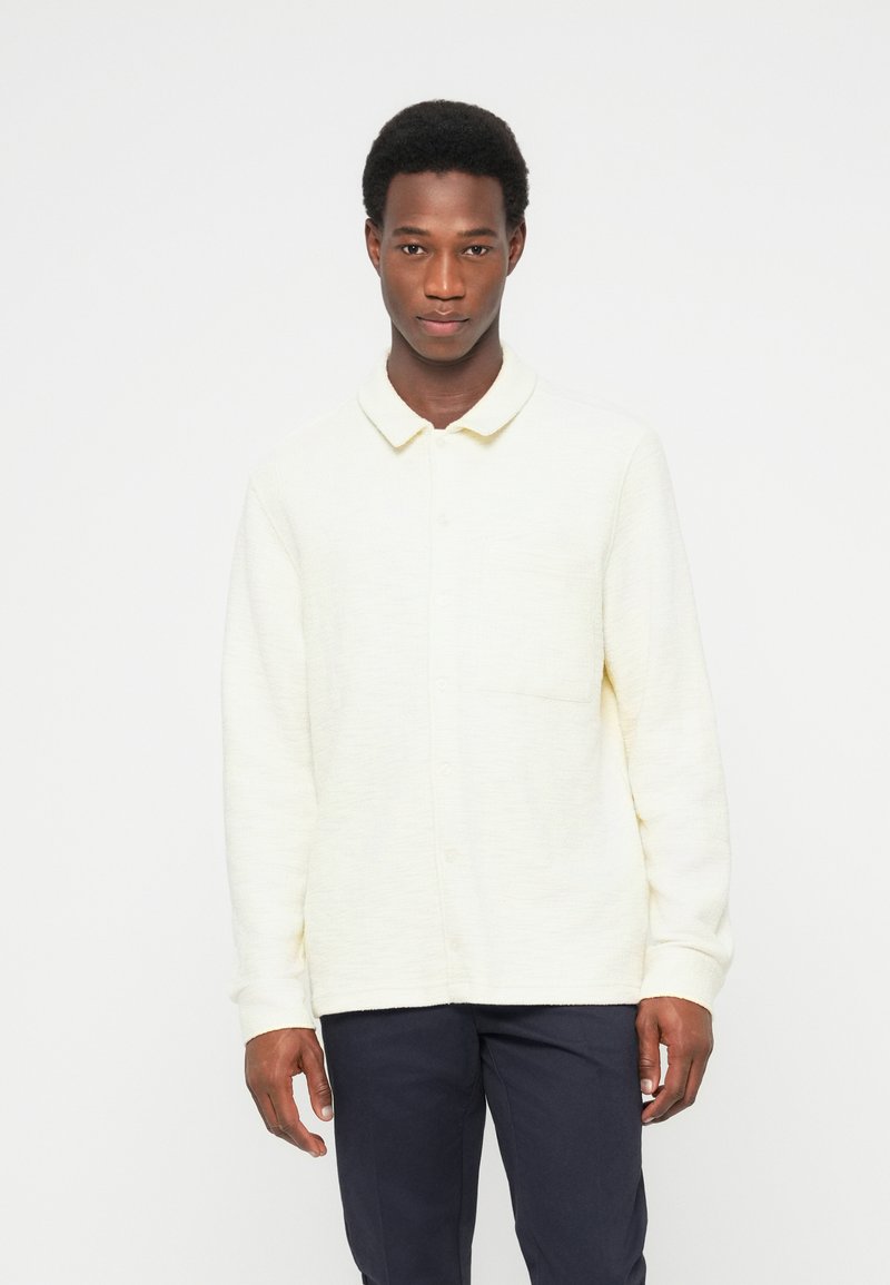 Ανοιχτόκίτρινο πουκάμισο με κουμπιά από υφασμάτινη υφή, με γιακά και μία τσέπη στο στήθος, συνδυασμένο με σκούρο παντελόνι.