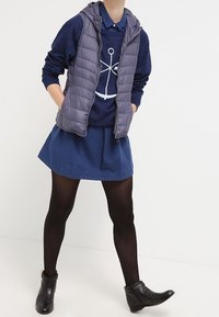 Gepolsterte graue Weste über einem marineblauen Pullover mit grafic in Weiß, kombiniert mit einem blauen Jeansrock und schwarzen Stiefeletten. Schwarze Strumpfhosen runden den Look ab.