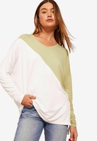 Majica s dolgimi rokavi z diagonalnim razporedom v svetlo zeleni in beli barvi, z dekoltejem v obliki črke V in sproščenim krojem, v kombinaciji z jeans hlačami.