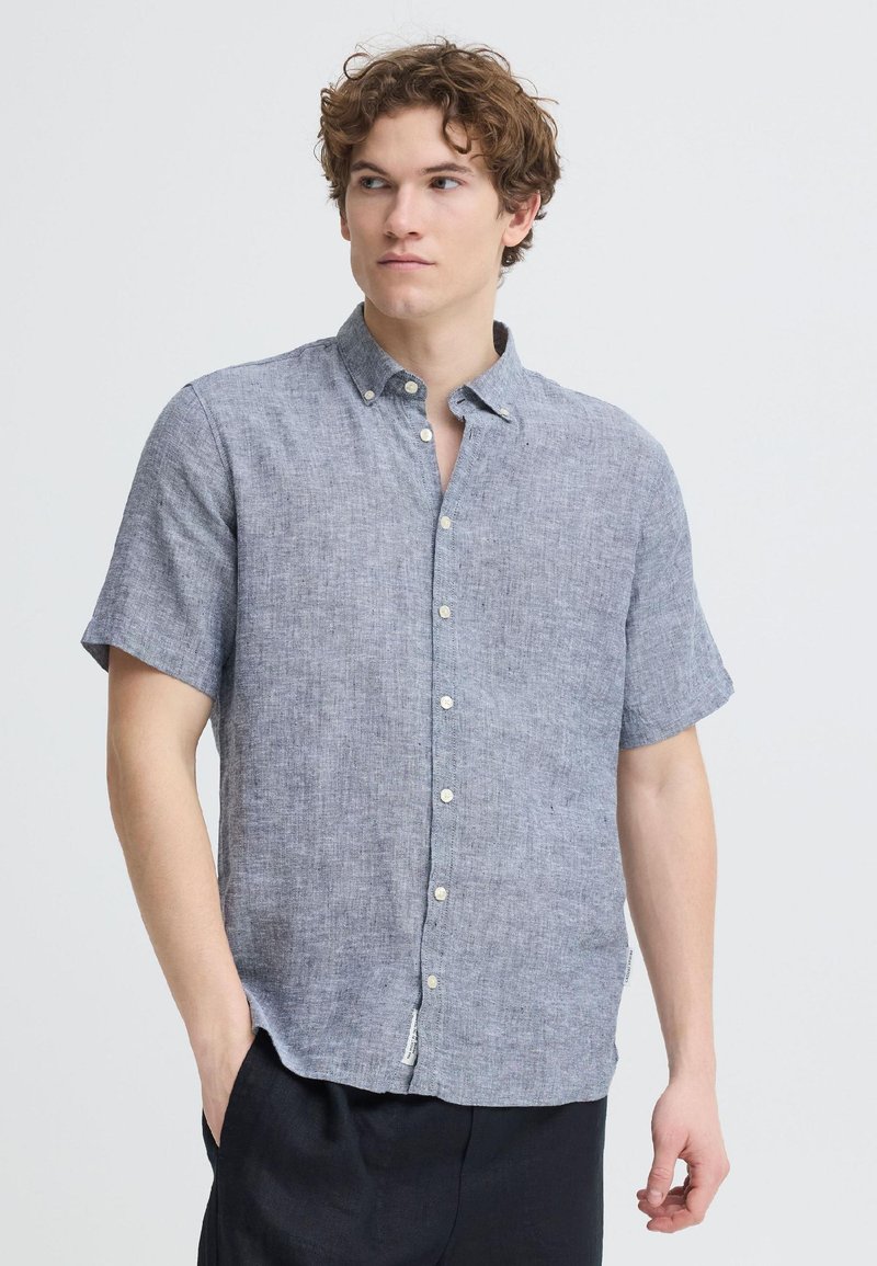 Jeune homme aux cheveux bouclés portant une chemise en lin bleu-gris à manches courtes boutonnée et un pantalon noir, regardant sur le côté avec la main dans la poche.