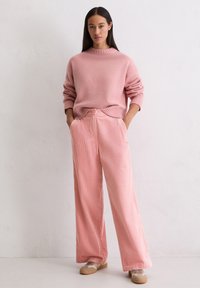 Jasnoróżowy, oversize'owy sweter z prążkowanym dekoltem i mankietami, w połączeniu z jasnoróżowymi sztruksowymi spodniami o szerokich nogawkach. Obuwie z beżowymi akcentami.