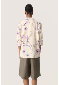 Helle beigen Bluse mit lila floralem Muster, langen Ärmeln und Kragen, kombiniert mit olivgrünen, knielangen Shorts. Glattes Material.