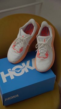 Zapatillas deportivas blancas con acentos naranjas, parte superior de malla y plantillas con motivo, descansando sobre una caja de zapatos HOKA azul contra un fondo amarillo.