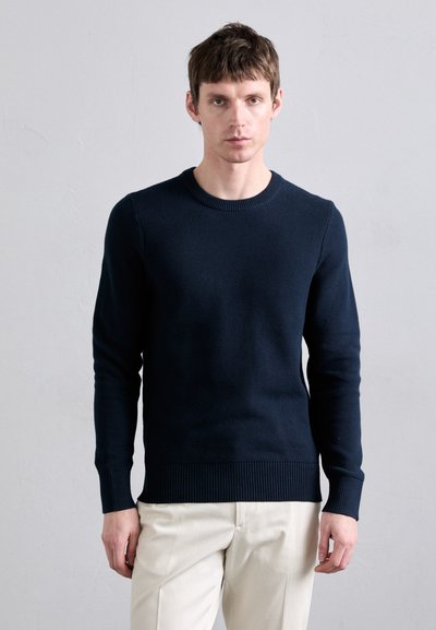 Pullover lavorato a maglia blu navy con scollatura a girocollo, polsini e orlo a coste. Il tessuto presenta un motivo strutturato, abbinato a pantaloni di colore chiaro.