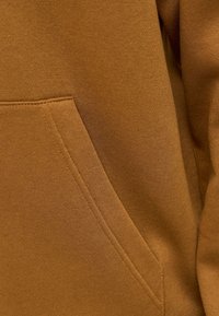 Barna szövet, melyen közeli kép látható egy átlós varrásról és zsebvarrásról egy ruhadarabon, valószínűleg egy pulóveren vagy kapucnis felsőn.