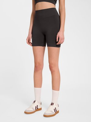 Kvinde iført sorte højttaljede biker-shorts, hvide crew-sokker og hvide sneakers med brune detaljer, stående på en ensfarvet lys baggrund.