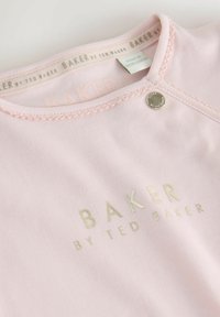 Lyserød bomuldsbluse med rund halsudskæring, prydet med guldpræget tekst "BAKER by TED BAKER" og en sølvtrykknapdetalje ved skulderen.