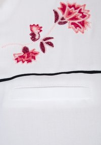 Bílá tkanina s malou našitou kapsou. Obsahuje růžovou květinovou výšivku s červenými a tmavě vínovými akcenty. Černá linka nad kapsou.