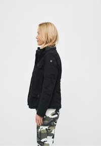 Giacca in denim nera con chiusura a bottoni e zip, con collo alto e due tasche frontali. Pantaloni con stampa camouflage nelle tonalità di verde e beige.