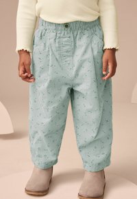 Helle grüne Hose aus strukturiertem Stoff mit floralem Muster, ausgestattet mit einem elastischen Bund und Seitentaschen, kombiniert mit grauen Knöchelstiefeln.